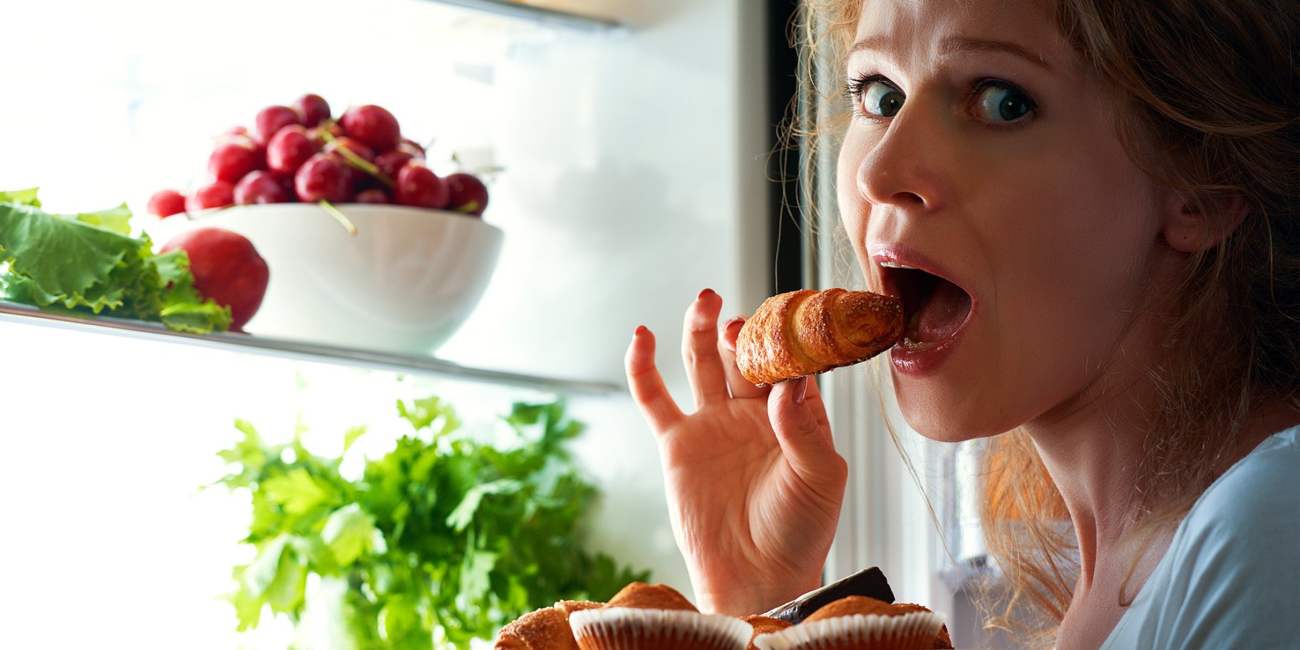Жінка їсть круасан біля холодильника